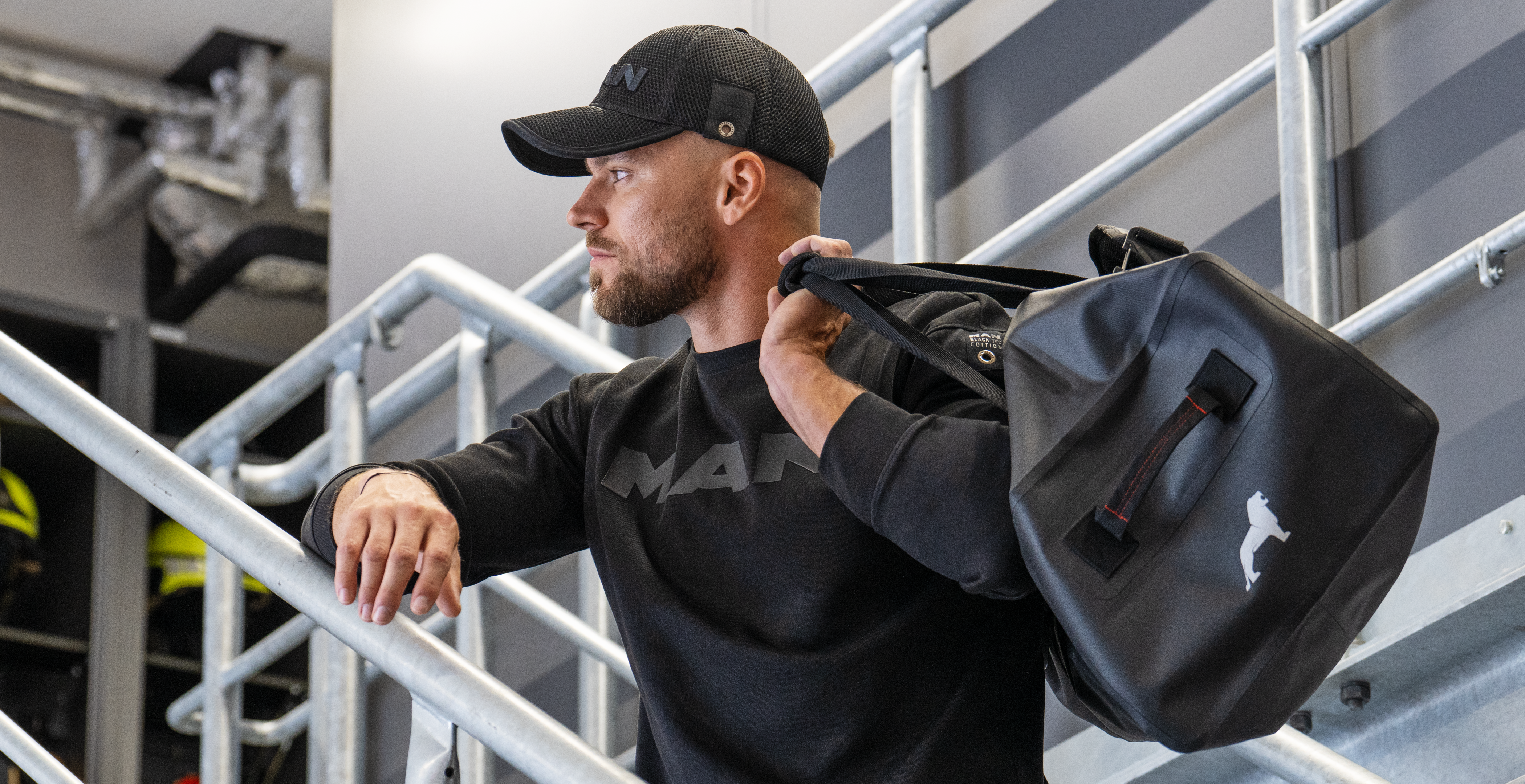 Mann in schwarzem MAN-Pullover und schwarzer Cap mit stylischer Tasche über der Schulter, ideal für einen modernen, aktiven Lebensstil.