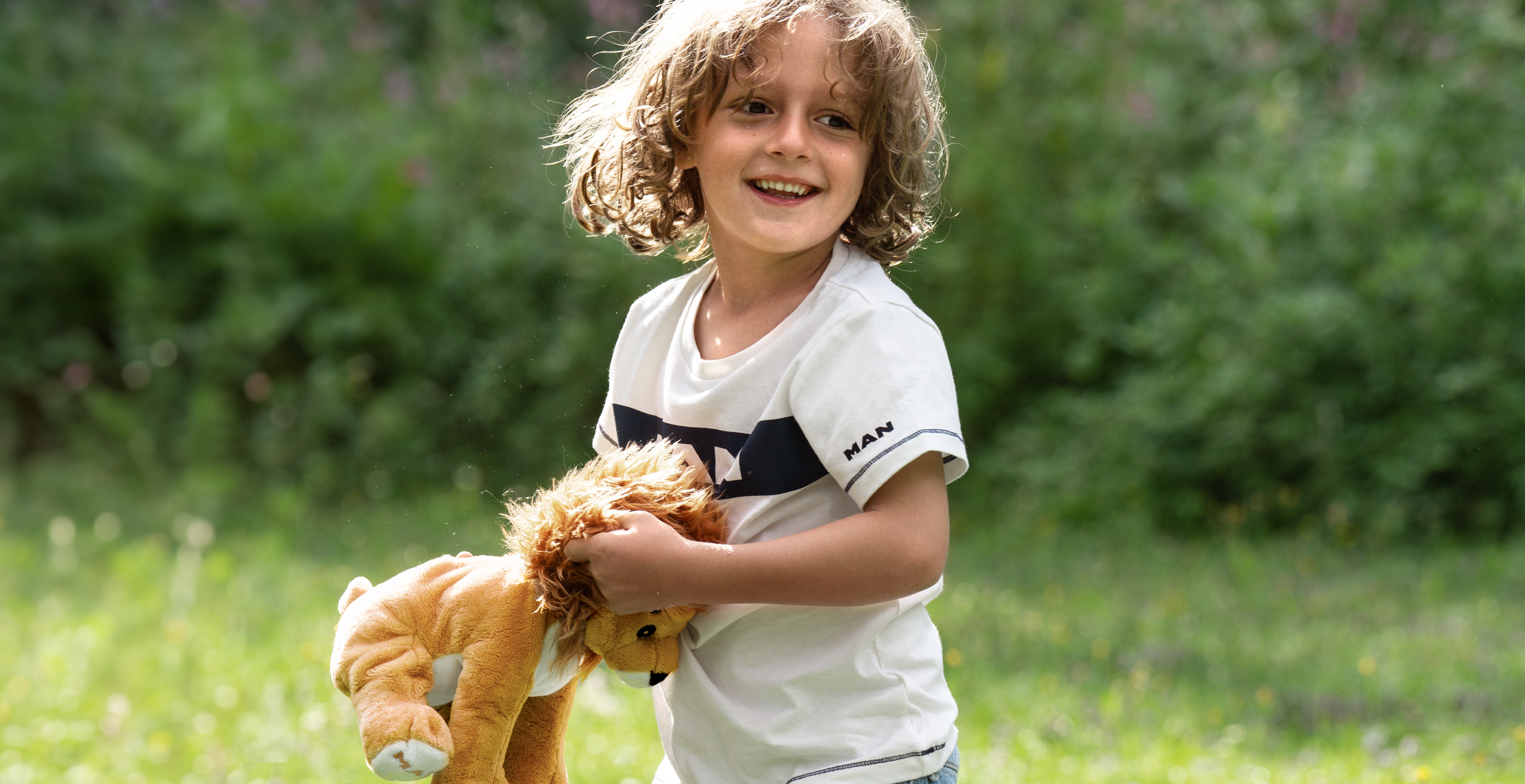 Lächelndes Kind in einem MAN-T-Shirt hält ein Löwen-Plüschtier, perfekt für aktive Kinder, die bequeme und stylische Kleidung lieben.