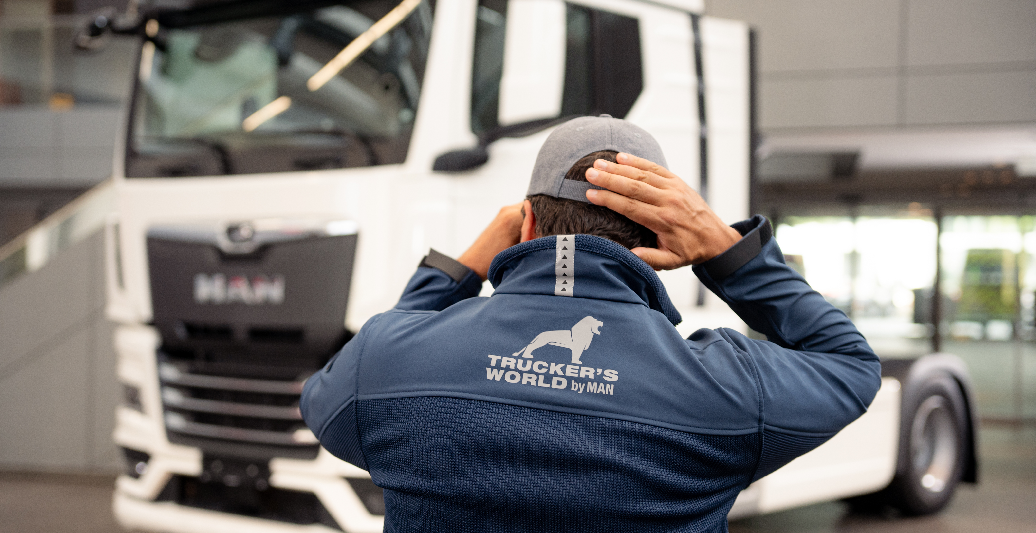 Trucker in blauer Trucker's World-Jacke und Cap, steht vor einem MAN-Lkw und passt seine Mütze an. Perfekte Kleidung für Trucker, die Stil und Funktionalität schätzen.